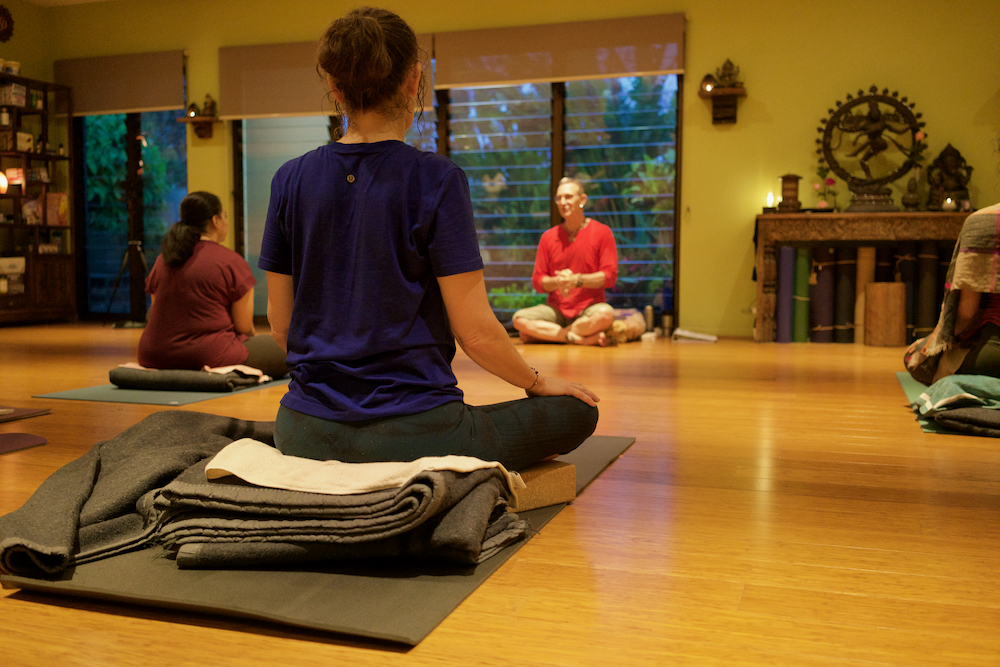 Yoga class in session with students seated on mats and blankets, listening to an instructor at the front of the studio.