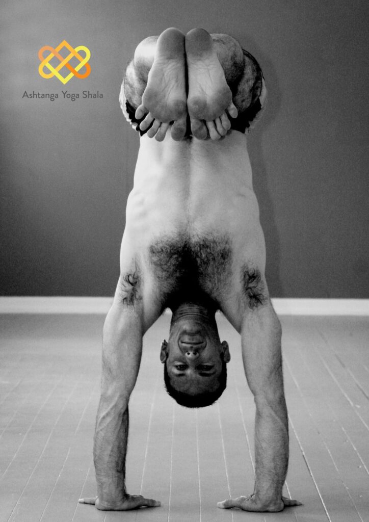 Black and white image of a yoga practitioner in a handstand variation, legs crossed behind the head.