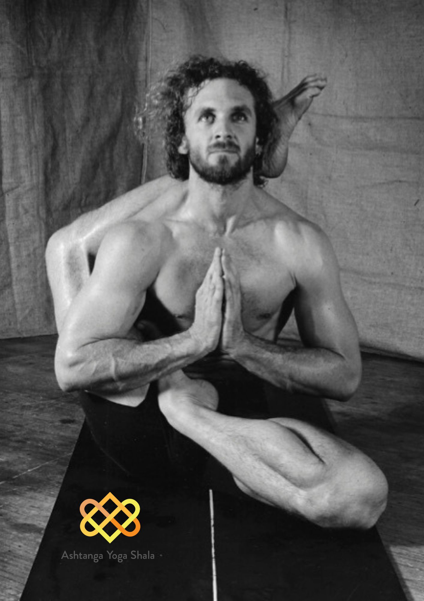 Black and white image of a man performing an advanced seated yoga posture with legs behind the head, hands in prayer.