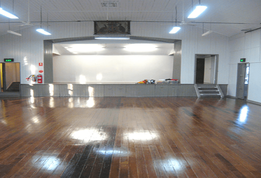 Large hall with dark timber floors, white walls, bright overhead lighting, and a small raised stage at the front.